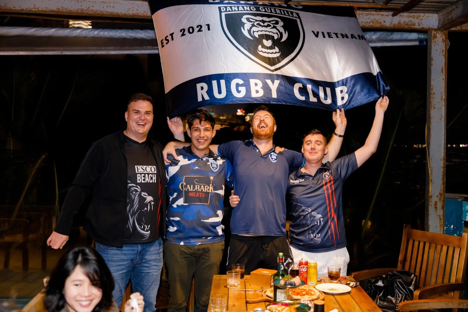 Four club members cheering under the Da Nang Guerilla Rugby Club flag